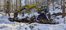 Vytváření meandrujícího koryta dočasného vodního toku. Bagrista Tomáš Homuta s využitím kráčejícího bagru přerušuje přímý odvodňovací příkop, vytváří základ meandrů a tůněk tak, aby se vodní tok v podmáčené rokli stal přírodě blízkým. Účinnost opatření vyhodnocují odborníci z Přírodovědecké fakulty Ostravské univerzity, Mgr. Stanislav Ruman, Ph.D. a RNDr. et Mgr. Matěj Horáček, Ph.D. Foto: Tomáš Salov